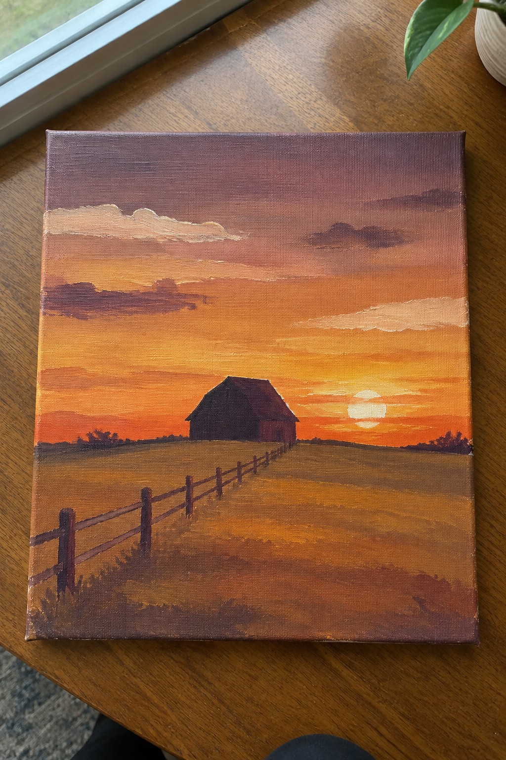 Painting of barn at sunset with wooden fence across golden field under orange sky