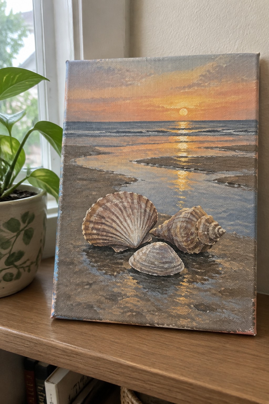 Seashell painting on canvas showing beach sunset scene placed beside potted plant on wood.