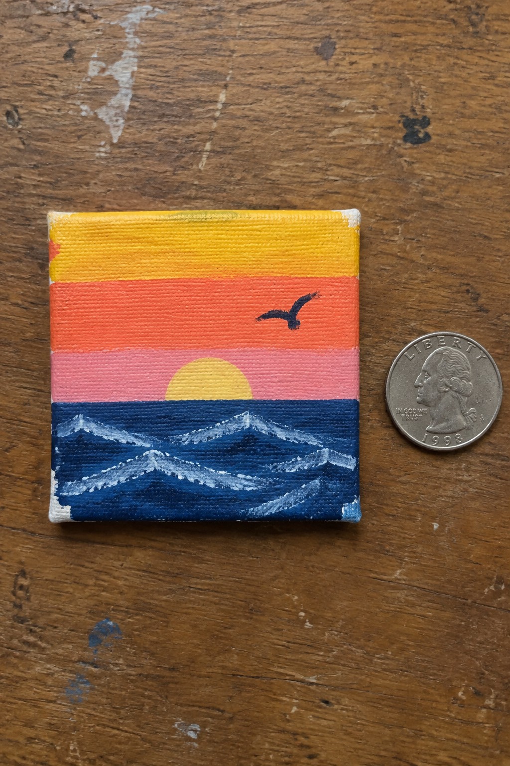 Miniature sunset painting of yellow-orange sky over blue waves beside quarter