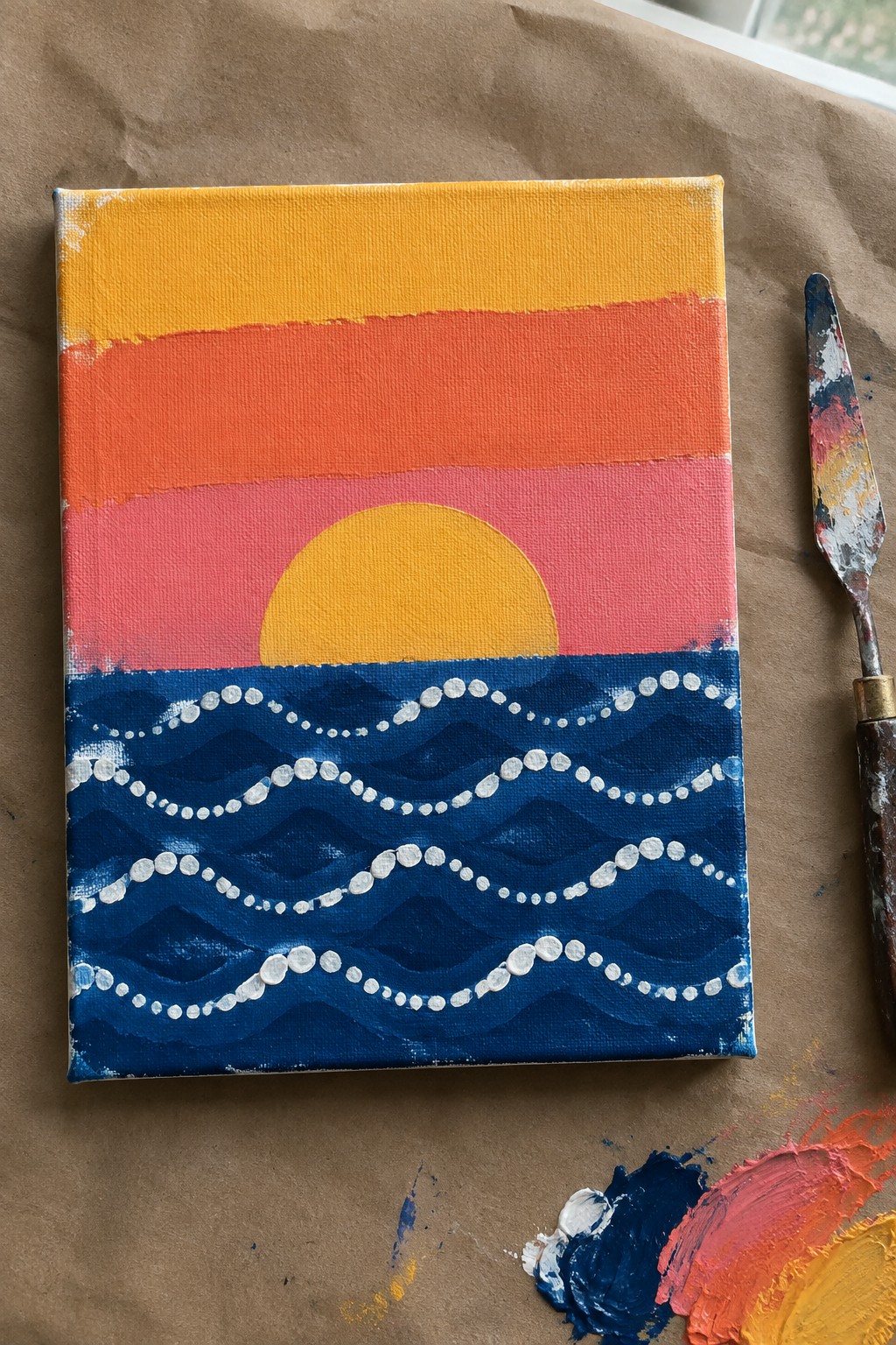 Sunset canvas painting with yellow sun over pink sky and dotted blue waves beside palette knife
