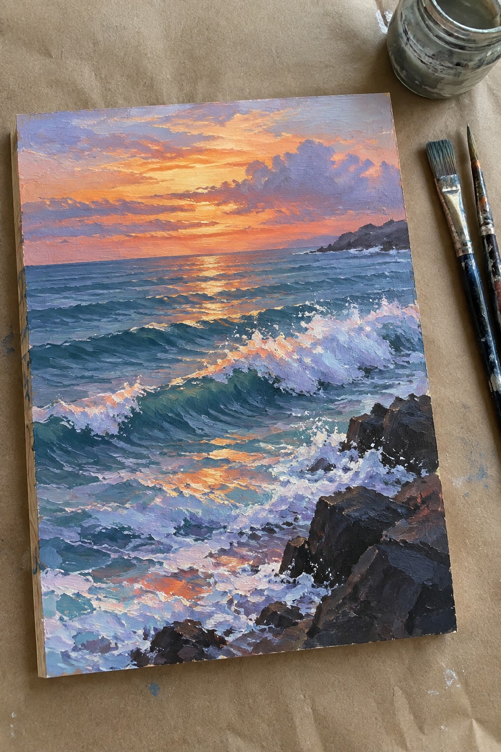 Vibrant oil painting of sunset waves crashing on rocks beside paintbrushes and jar