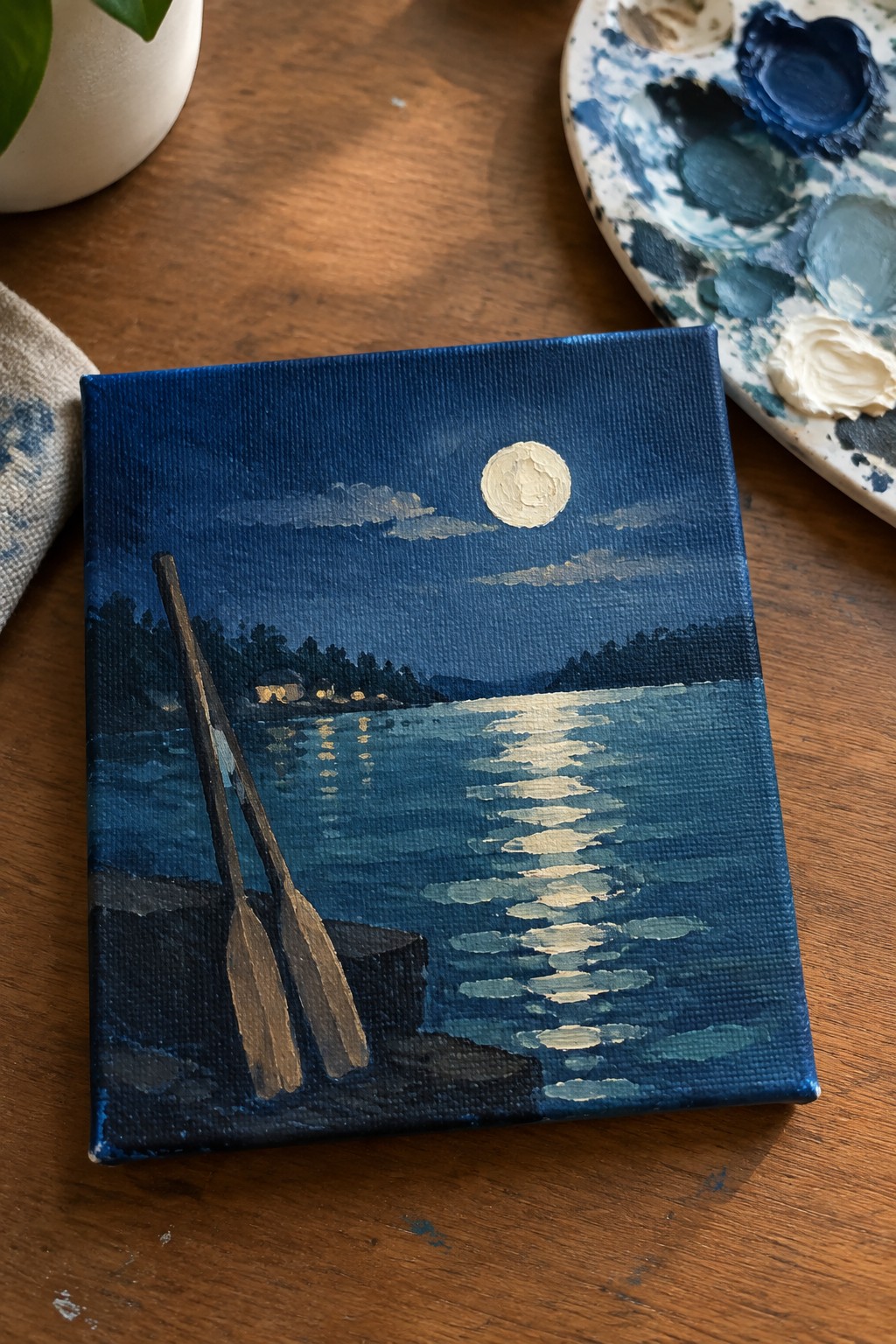 Canvas painting of moonlit lake with oars, palette on wooden table.