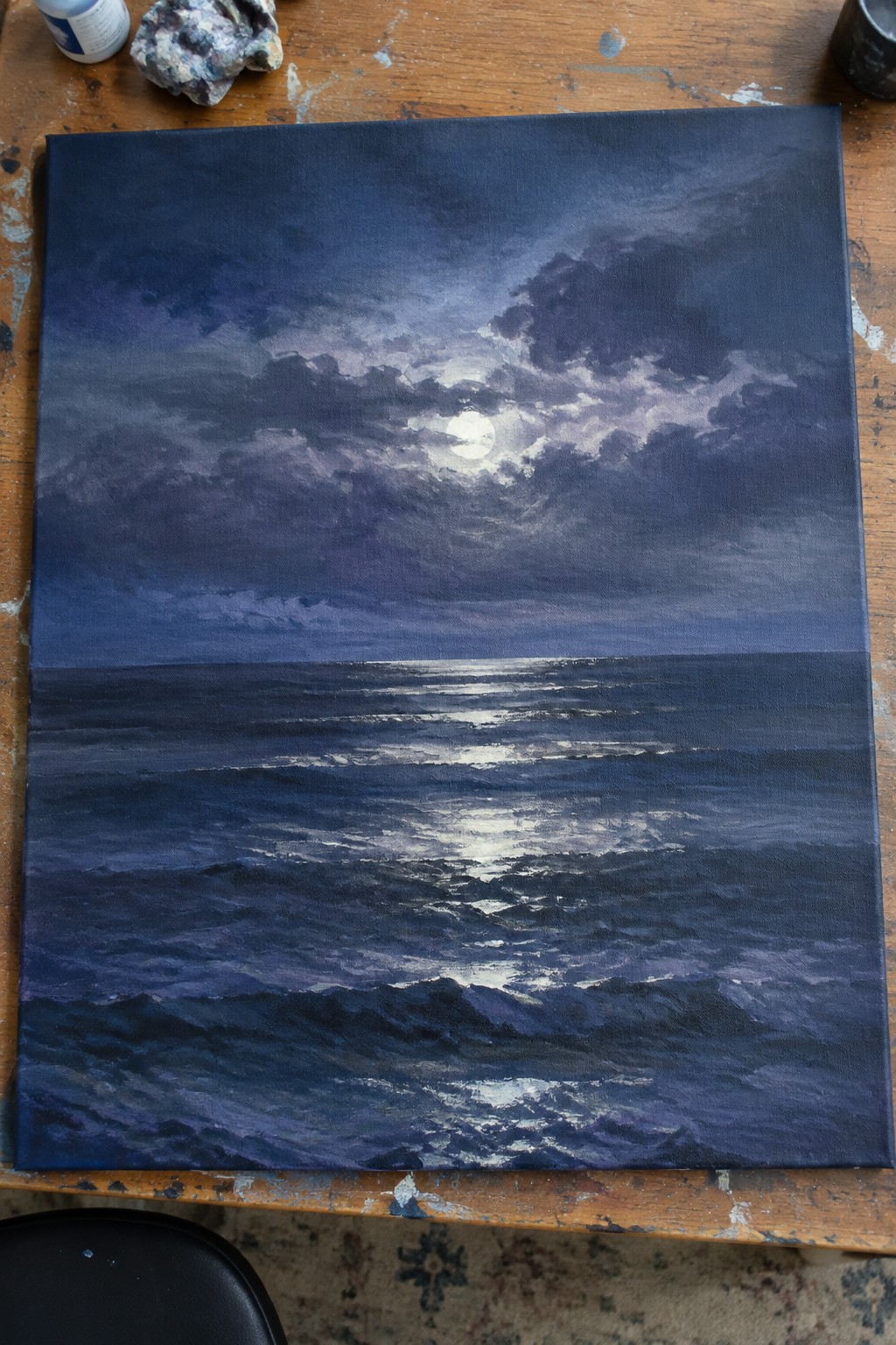 Moonlit stormy sea painting on canvas atop paint-splattered wooden table with supplies