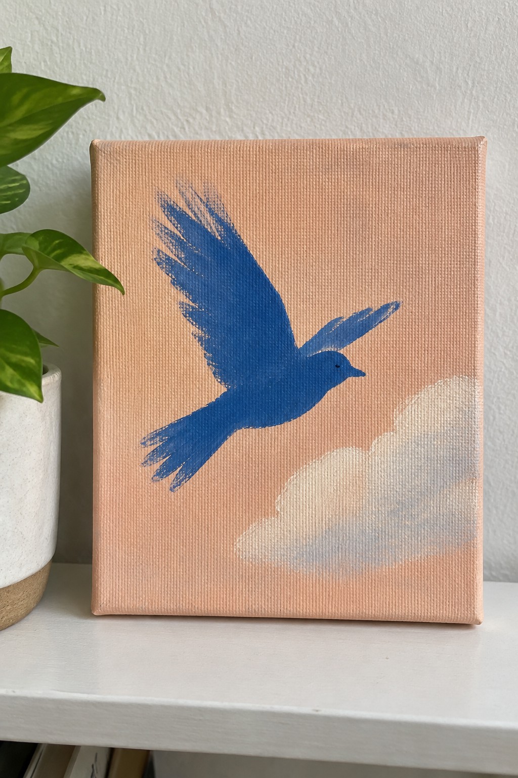 A blue bird flying across a peach-colored canvas with a white cloud in the lower right corner, next to a potted plant.