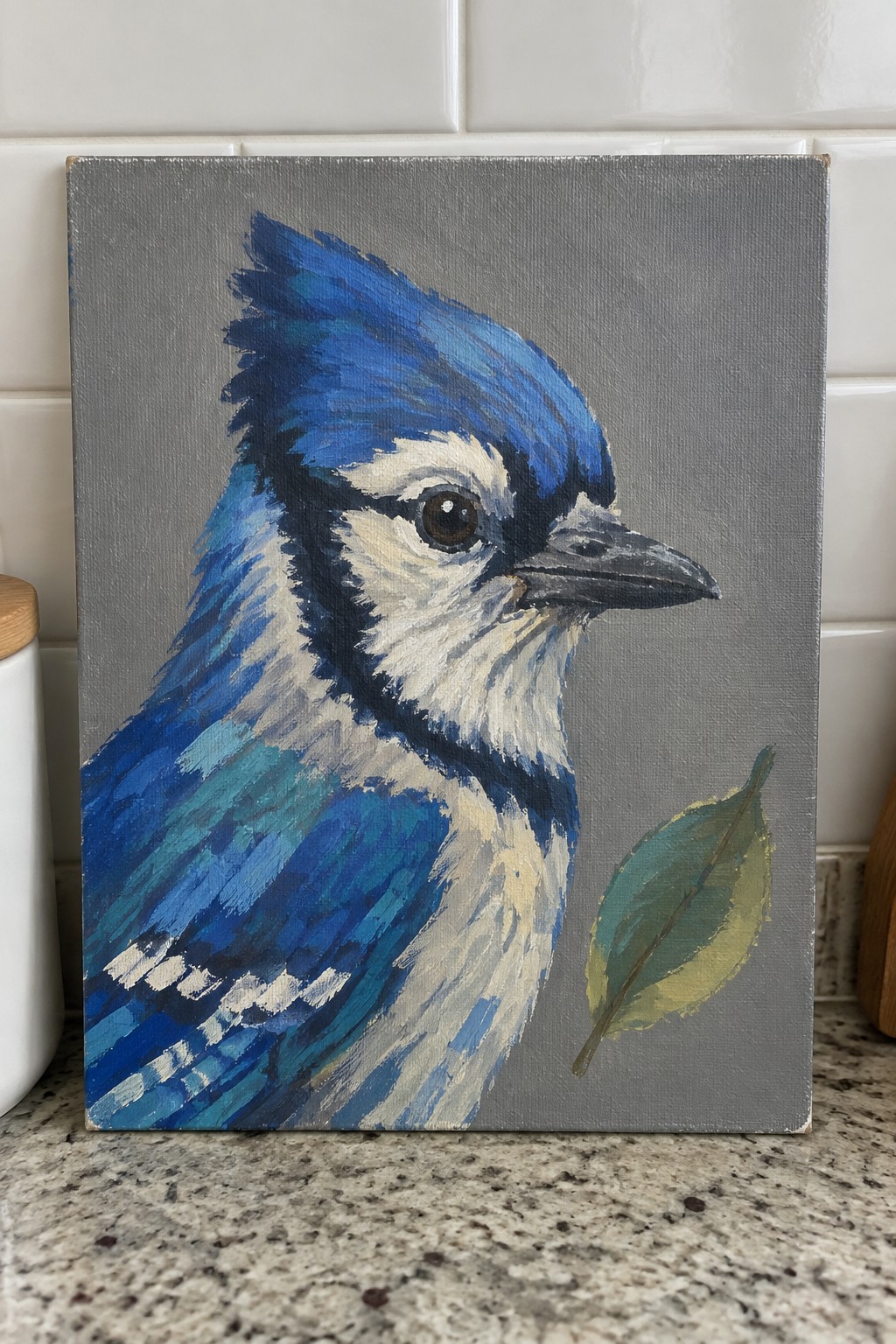 An acrylic painting of a blue jay in profile on a gray canvas with a small leaf painted in the lower right corner.