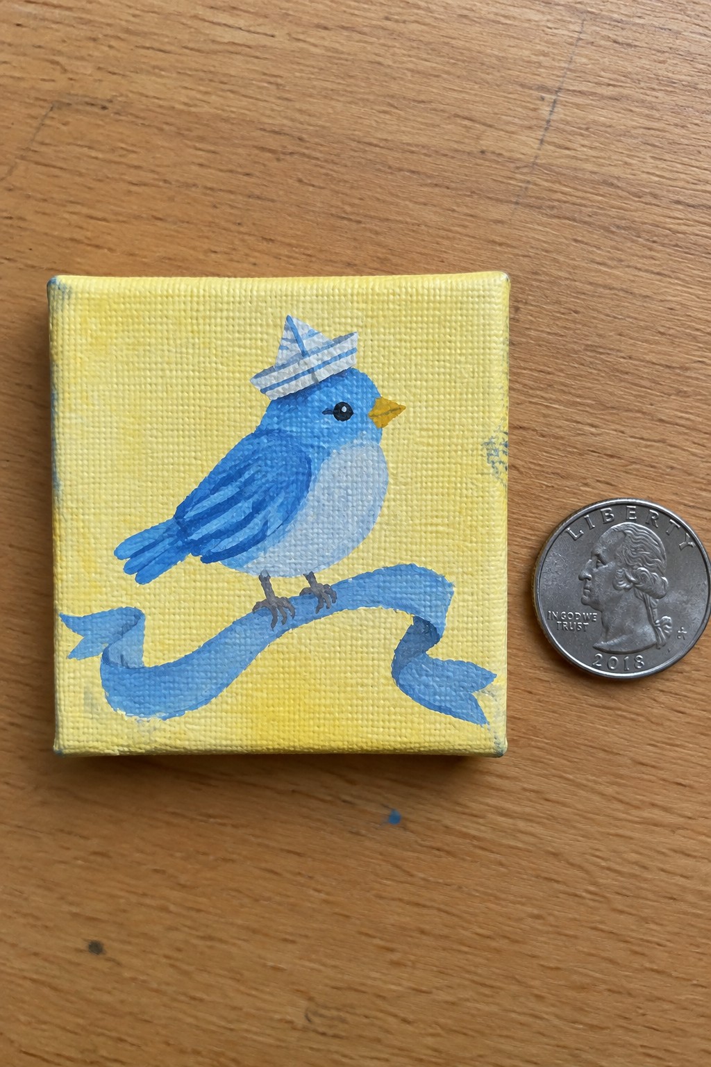 A small yellow canvas shows a blue bird wearing a striped paper hat perched on a blue ribbon, placed next to a 2018 quarter on a wooden surface.