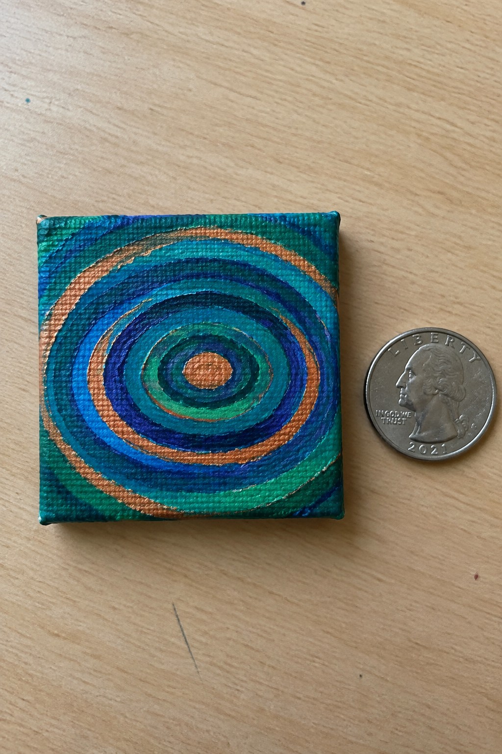 Small canvas with blue-green concentric circles beside a 2021 US quarter on wood.