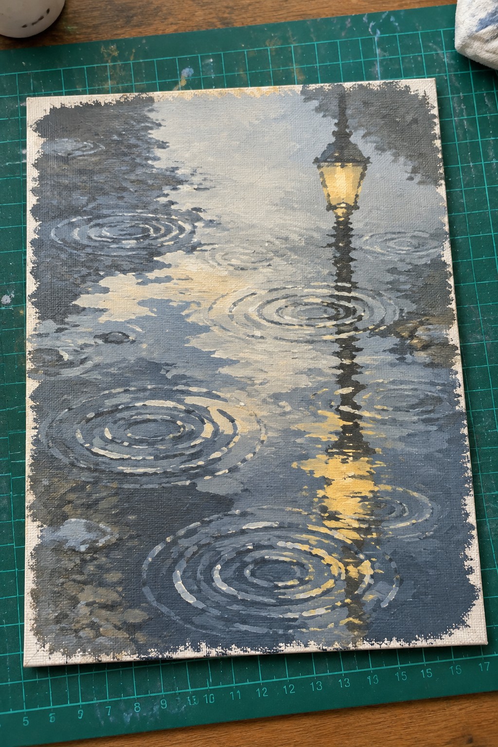 Painting of glowing streetlamp with yellow reflections in rippled rainy puddles