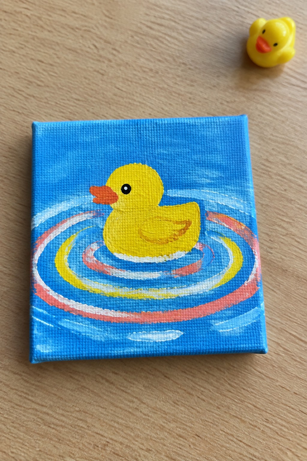 Yellow duck painting with colorful ripples beside small toy duck on wood