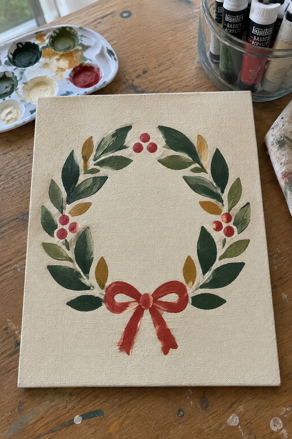 Painted canvas wreath with green leaves, red berries, and bow amid art supplies
