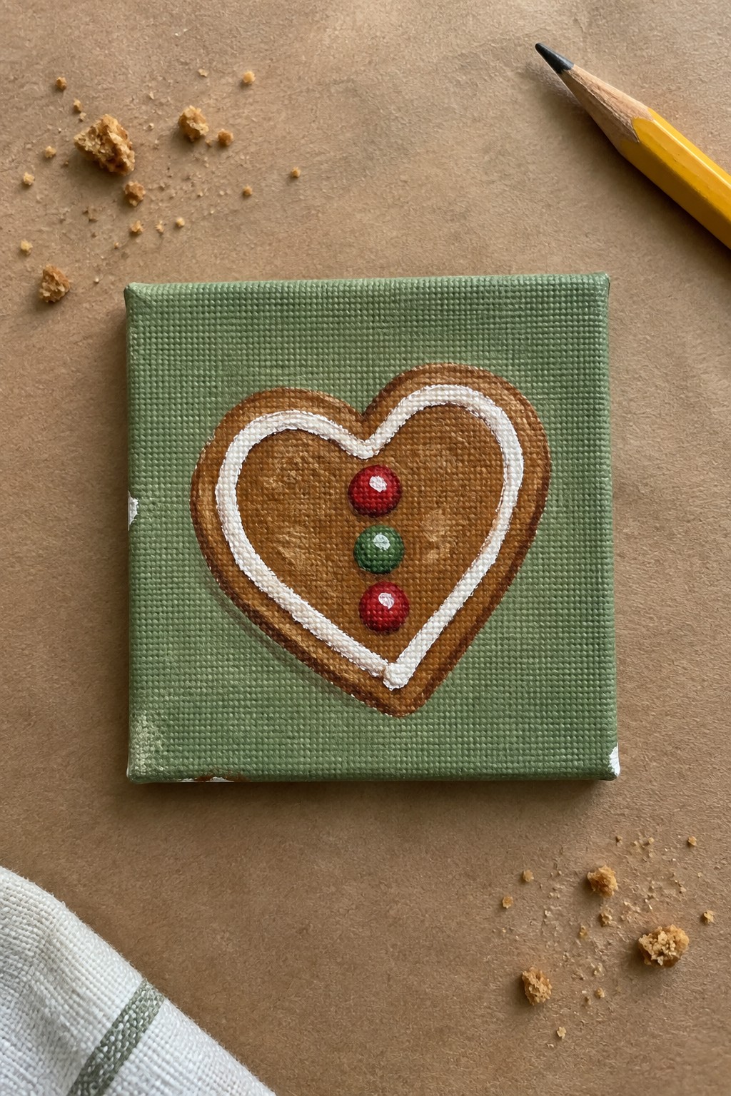 Heart-shaped gingerbread cookie painting on green canvas with yellow pencil and crumbs