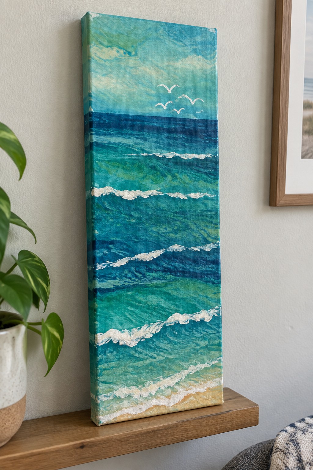 Turquoise ocean painting with waves, birds, and sand on wooden shelf beside plant
