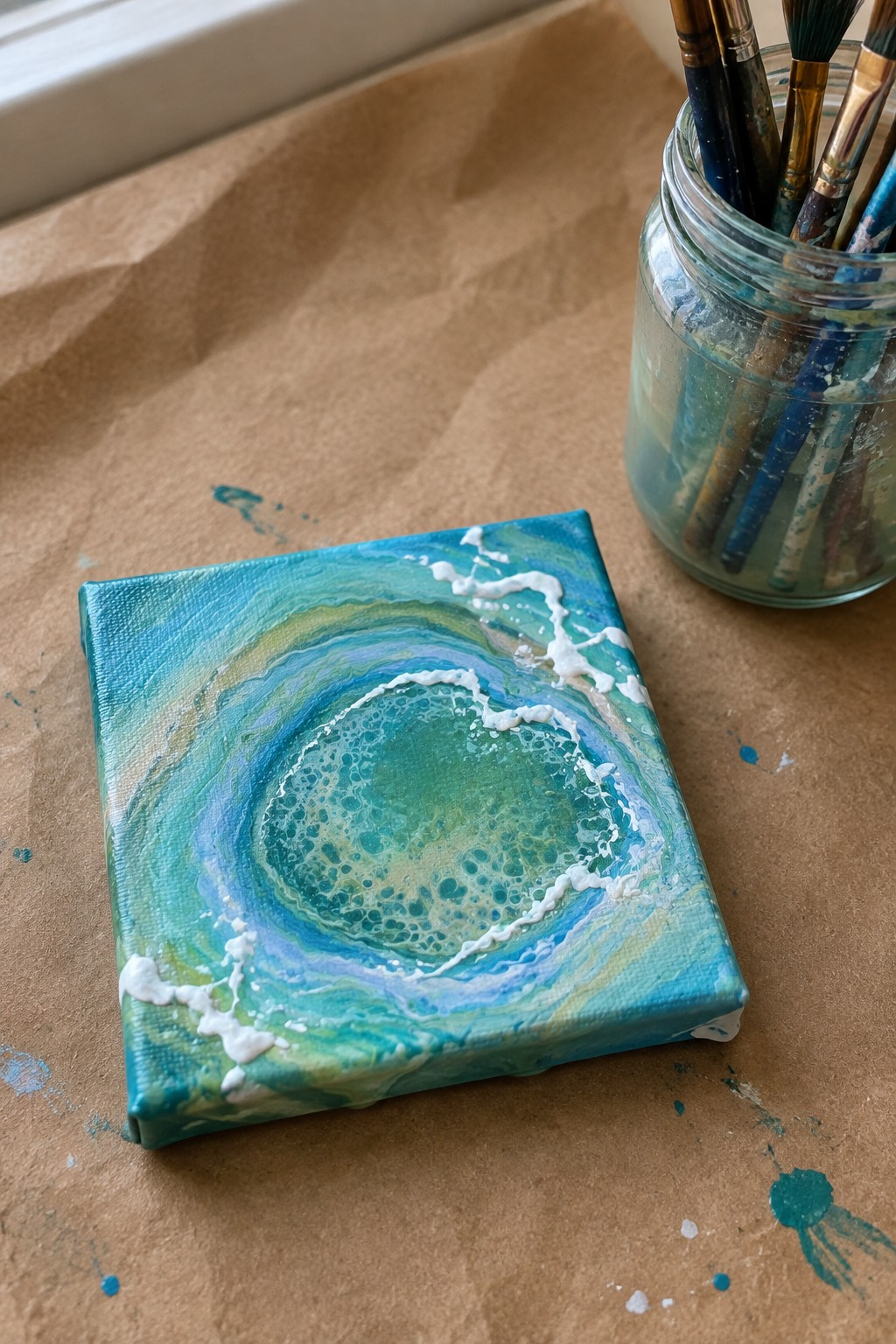 Mini canvas with swirling blue-green wave art beside jar of paintbrushes on paper.