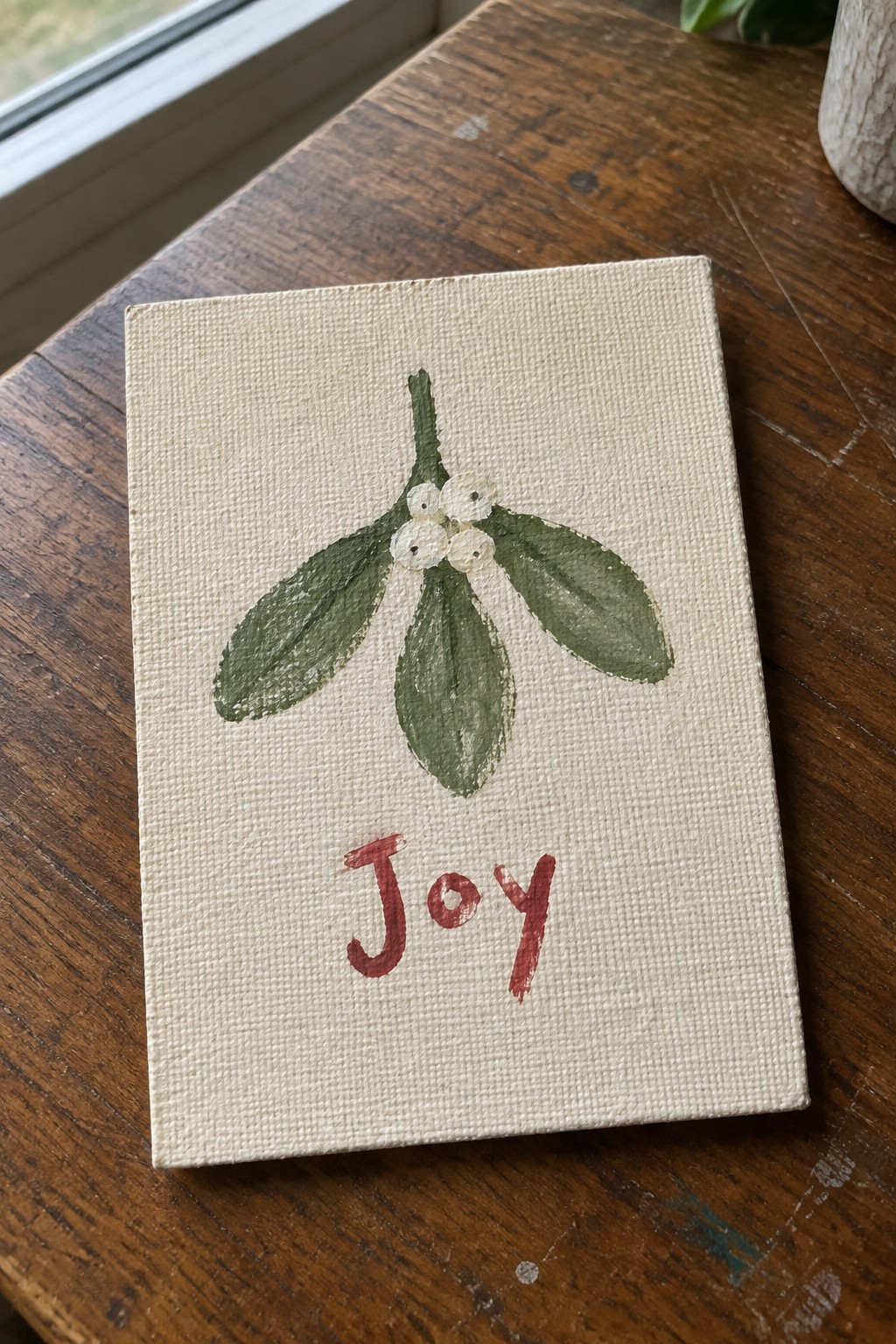 Beige canvas on wood shows green mistletoe with white berries above red 