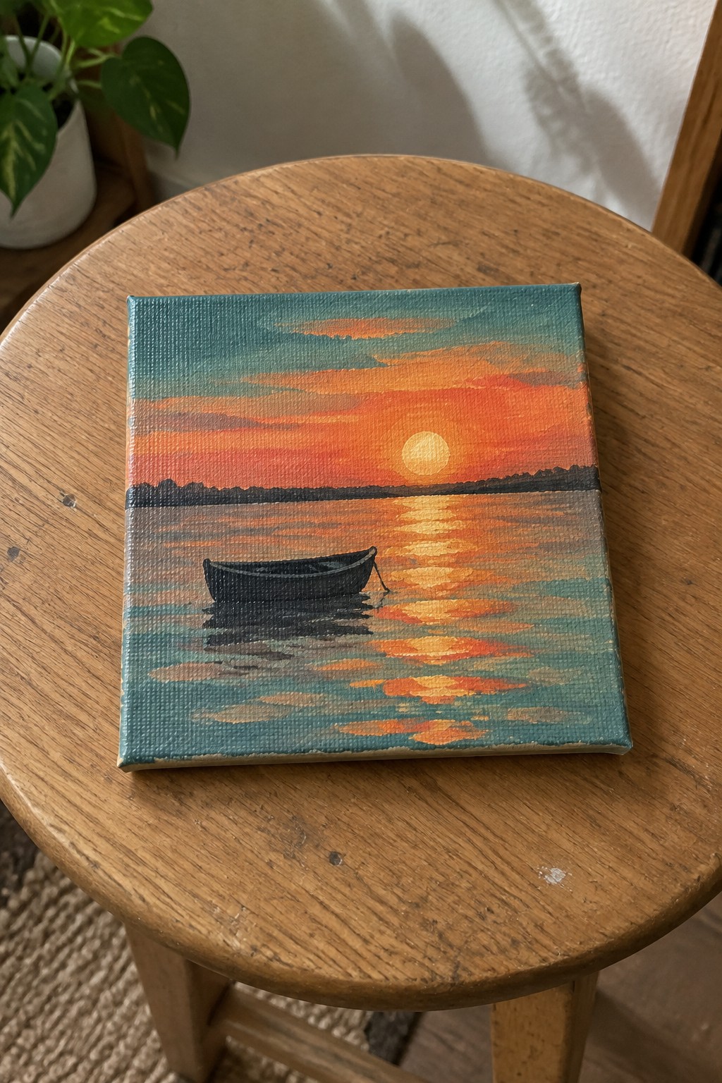 Small canvas sunset painting with boat on reflective water atop wooden stool.