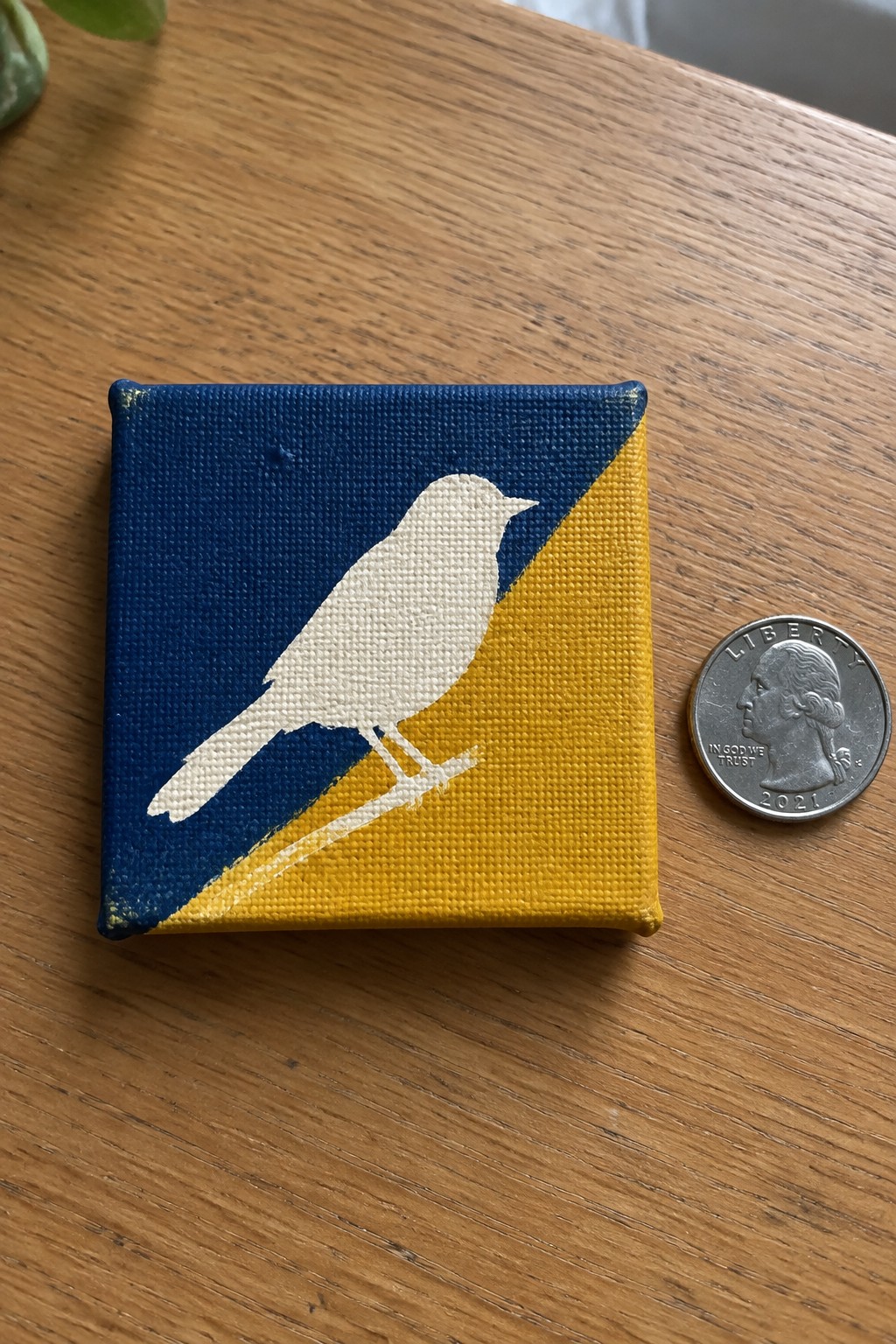 Small square canvas with white bird on blue-yellow diagonal beside quarter