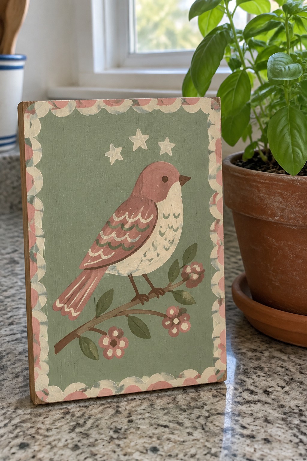 Folk art bird painting on scalloped wooden plaque beside potted basil plant on counter.