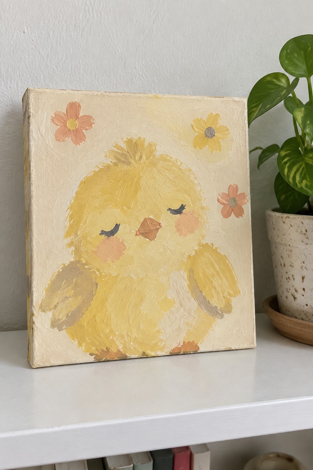 Textured painting of yellow chick with flowers on canvas beside potted plant on shelf