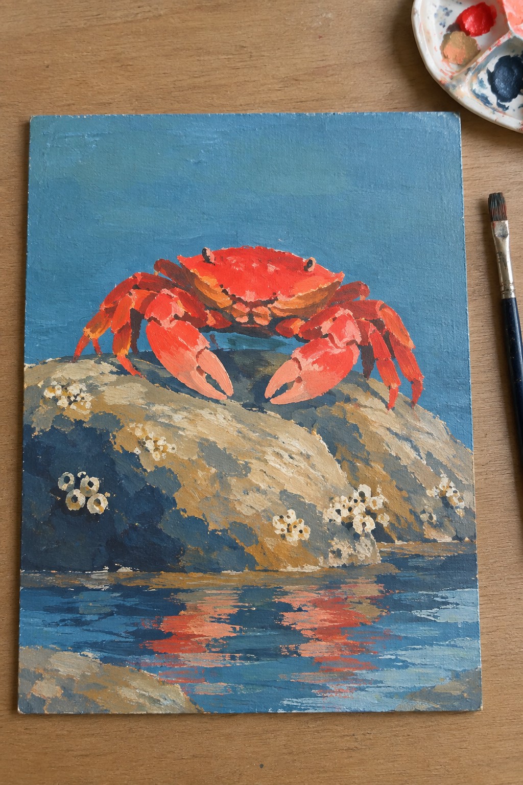 Vibrant red crab painting on canvas beside palette and brush on wooden table.