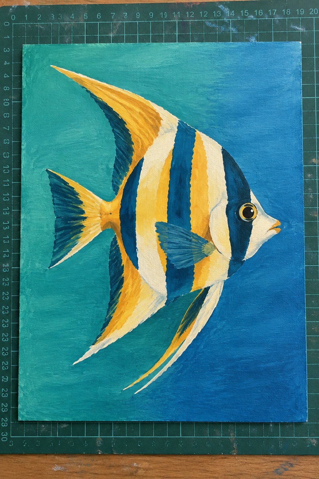 Striped yellow-blue fish painting on teal canvas atop green grid mat.
