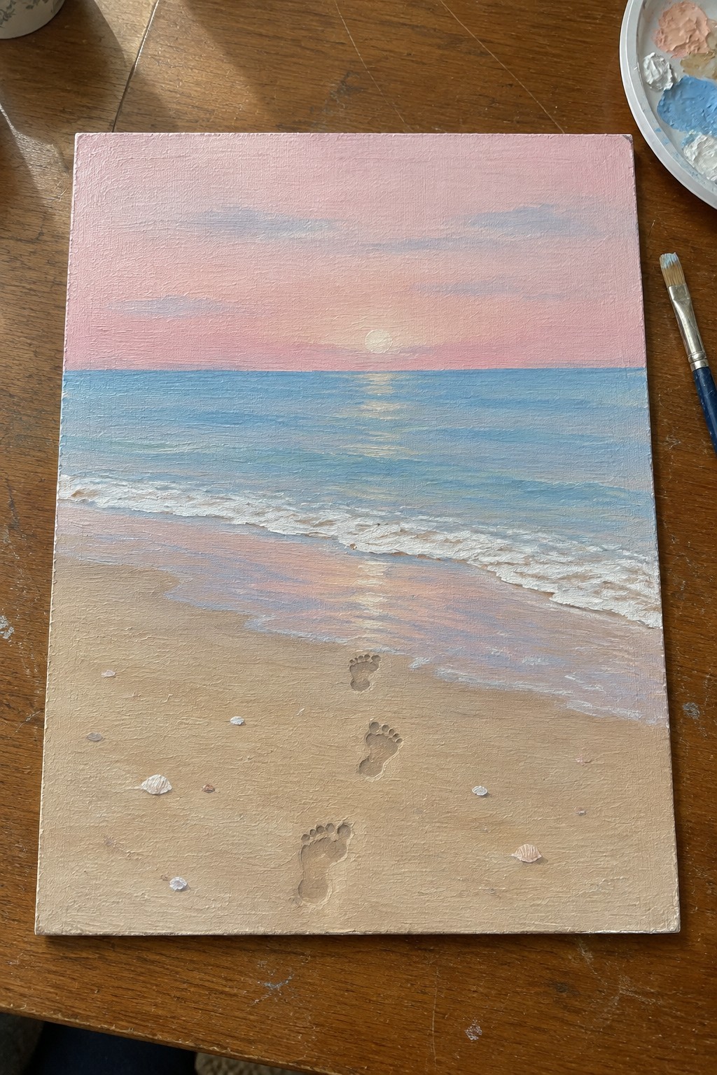 Beach sunset painting with footprints in sand, paintbrush and palette on table.