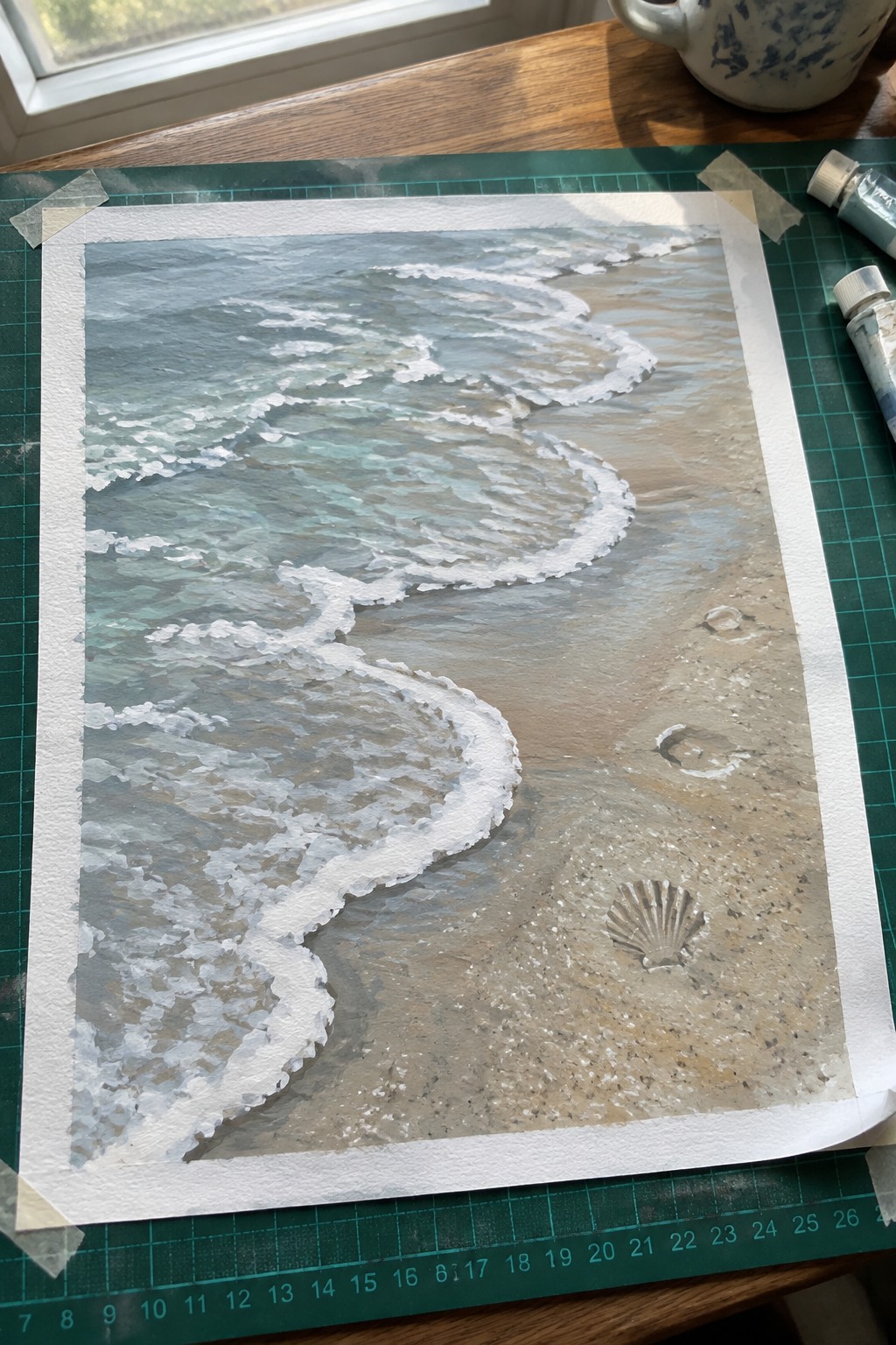 Watercolor painting of waves on sandy beach with seashell, taped to green cutting mat
