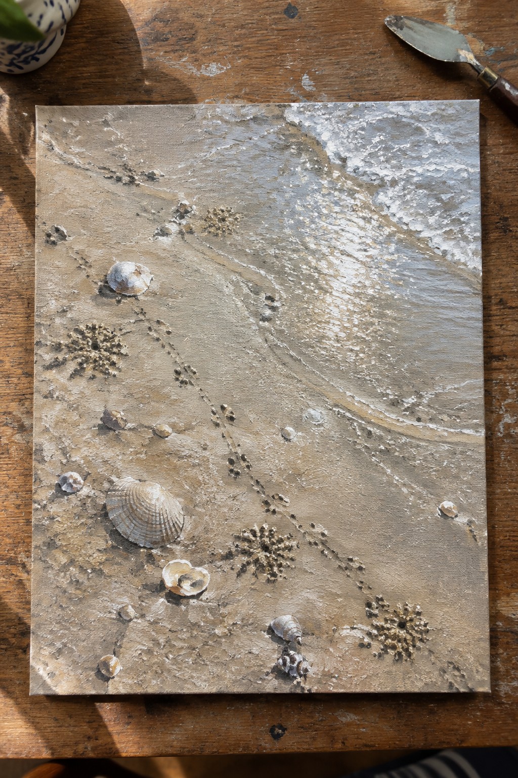 Thickly textured beach painting with waves, seashells, and footprints beside palette knife on wood