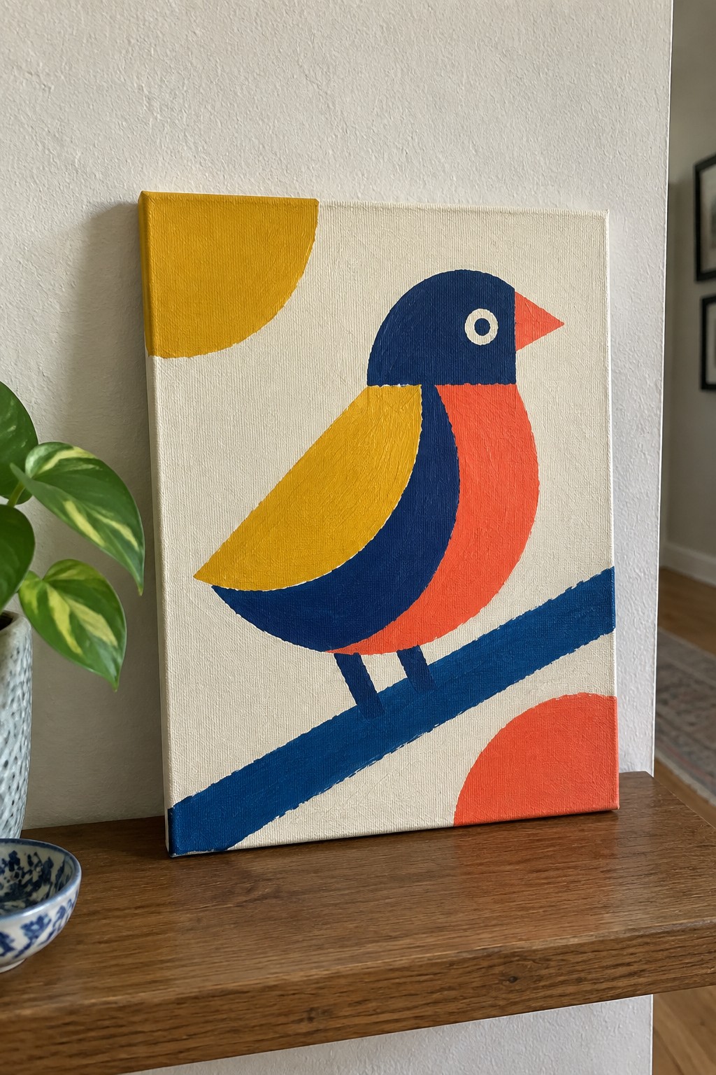 Abstract geometric bird painting with yellow wing, orange breast, blue head on wooden shelf