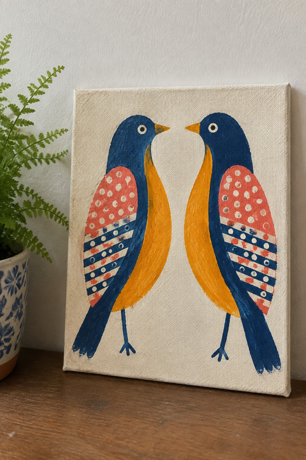 Two folk-art birds in blue, yellow, and red facing on a canvas beside a fern