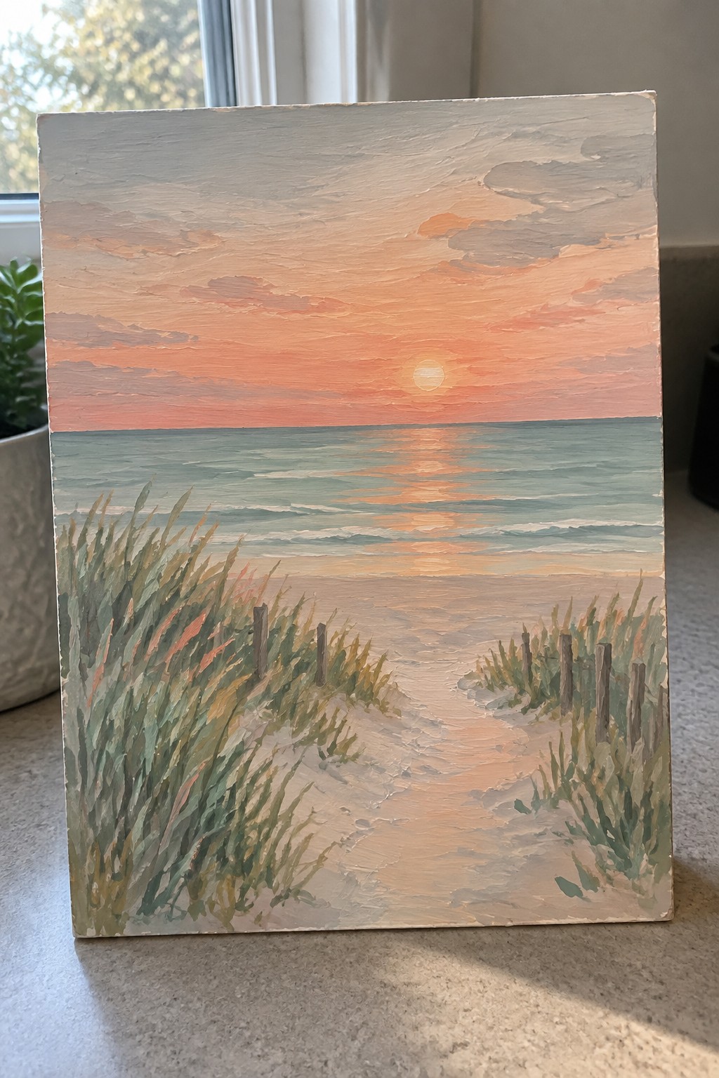 Acrylic painting of a beach sunrise with the sun low on the horizon, peachy reflections on the water, and a sandy path through grassy dunes.