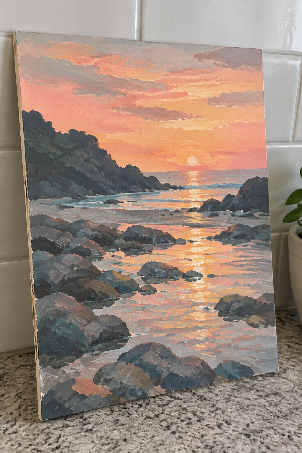 An acrylic painting of a sunset over the ocean with dark rocks in the foreground and peachy reflections on the water.