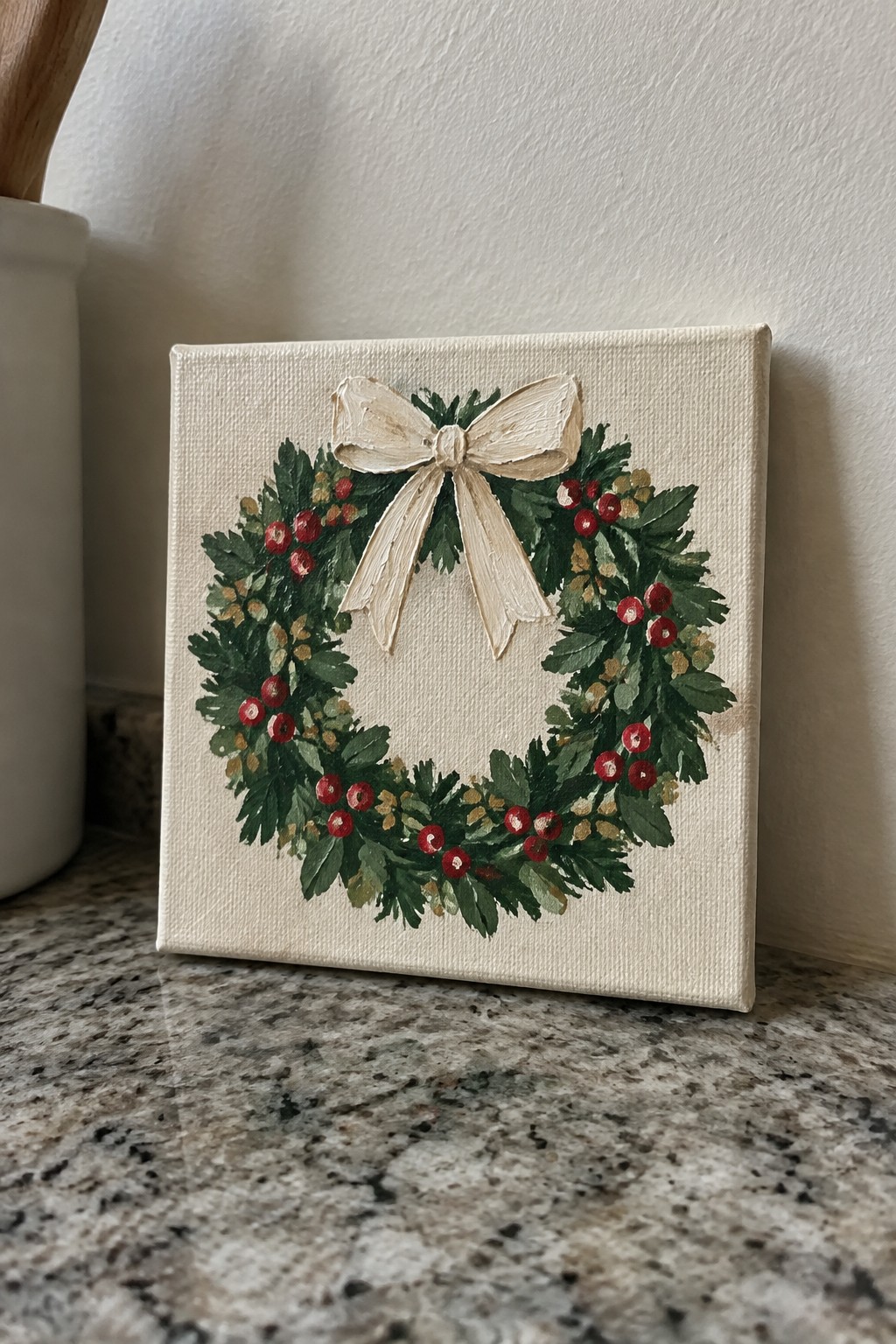 Canvas painting of festive wreath with green leaves, red berries, and beige bow on countertop
