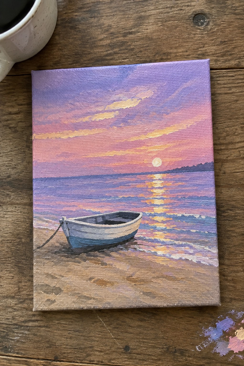 An acrylic painting of a small white and blue boat on a sandy beach at sunset with pink and purple skies and sun reflection on the water, sitting on a wooden table beside a mug and paint palette.