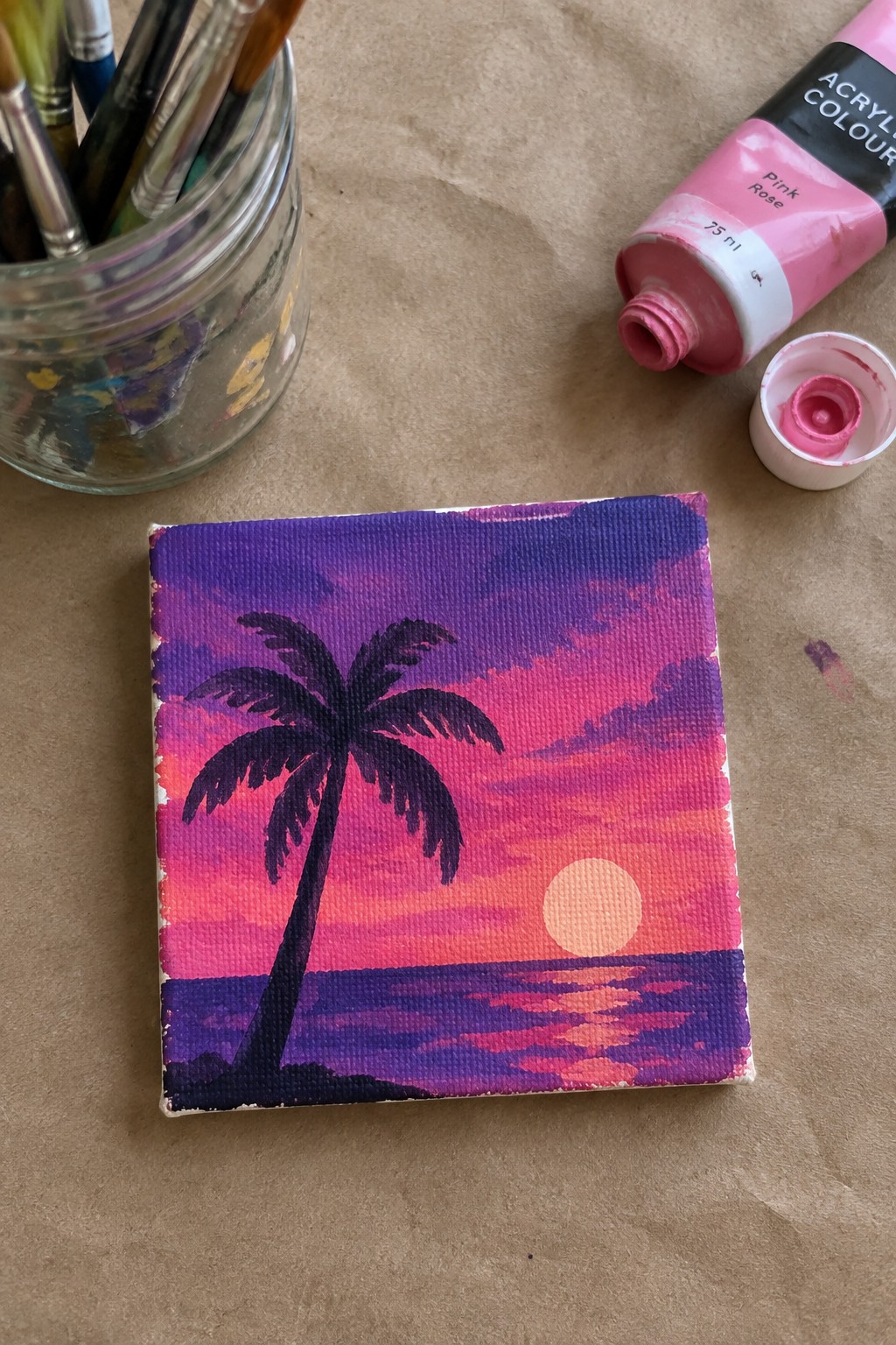 Mini canvas sunset painting with palm tree, paintbrushes, and open pink acrylic tube