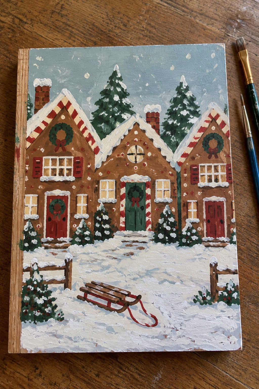 Snowy gingerbread houses with wreaths, red sled, and pine trees in winter scene.