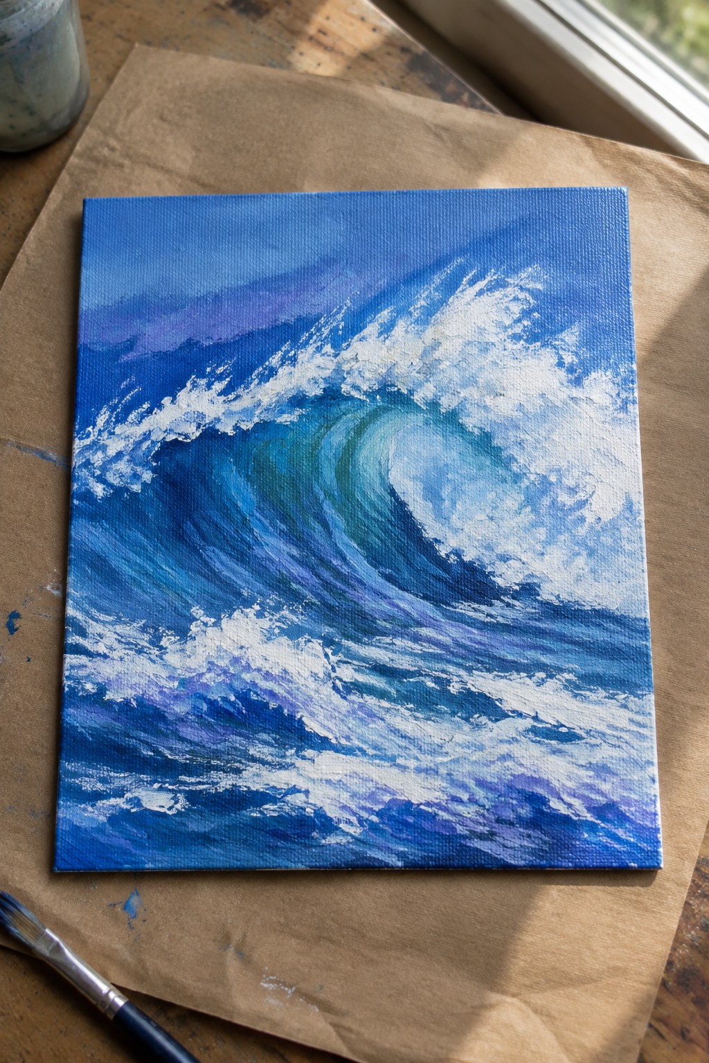 Canvas with blue wave painting on brown paper beside paintbrush on table.