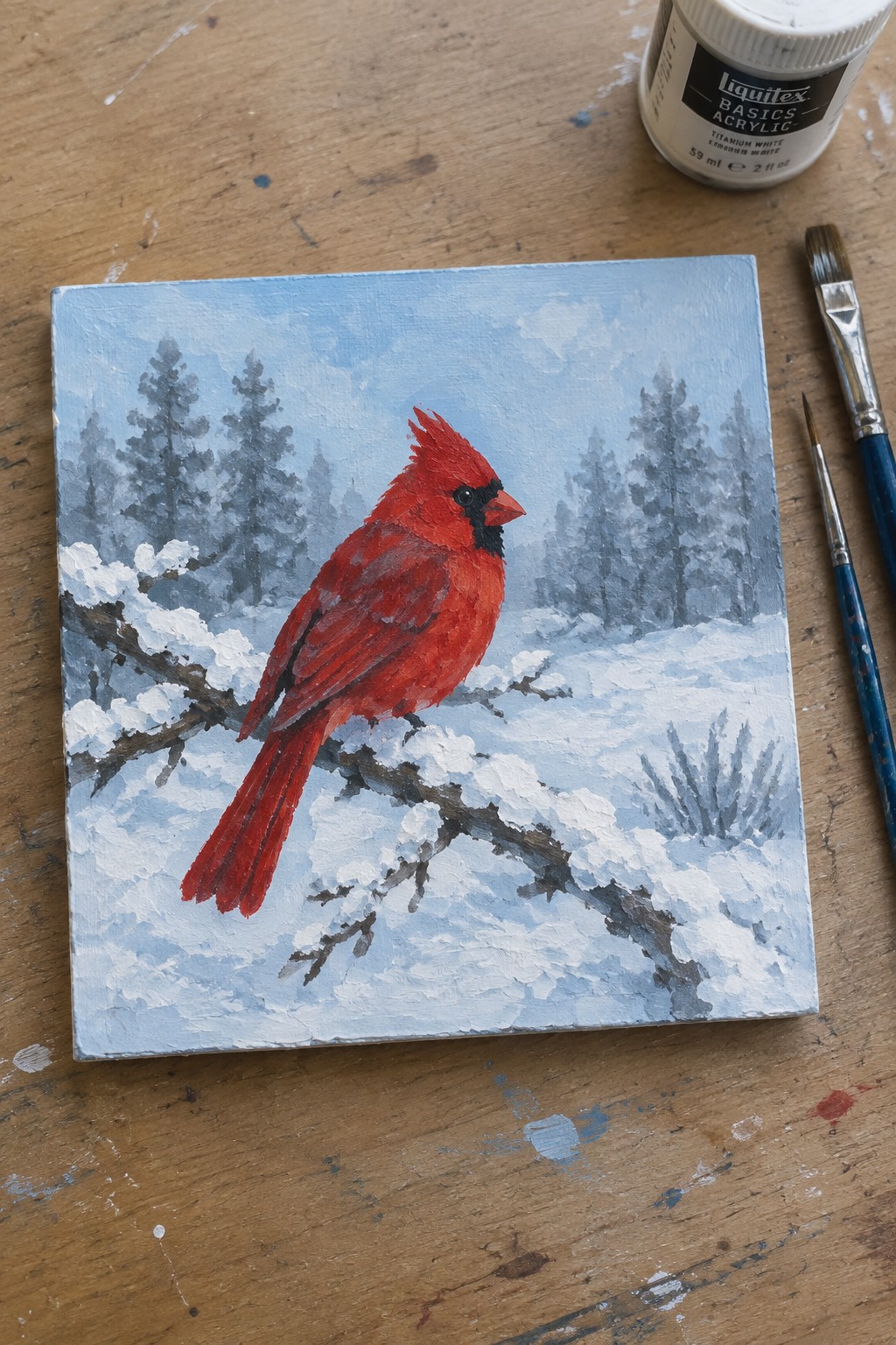 Red cardinal on snow-covered branch in acrylic winter painting, with brushes and paint tube nearby.