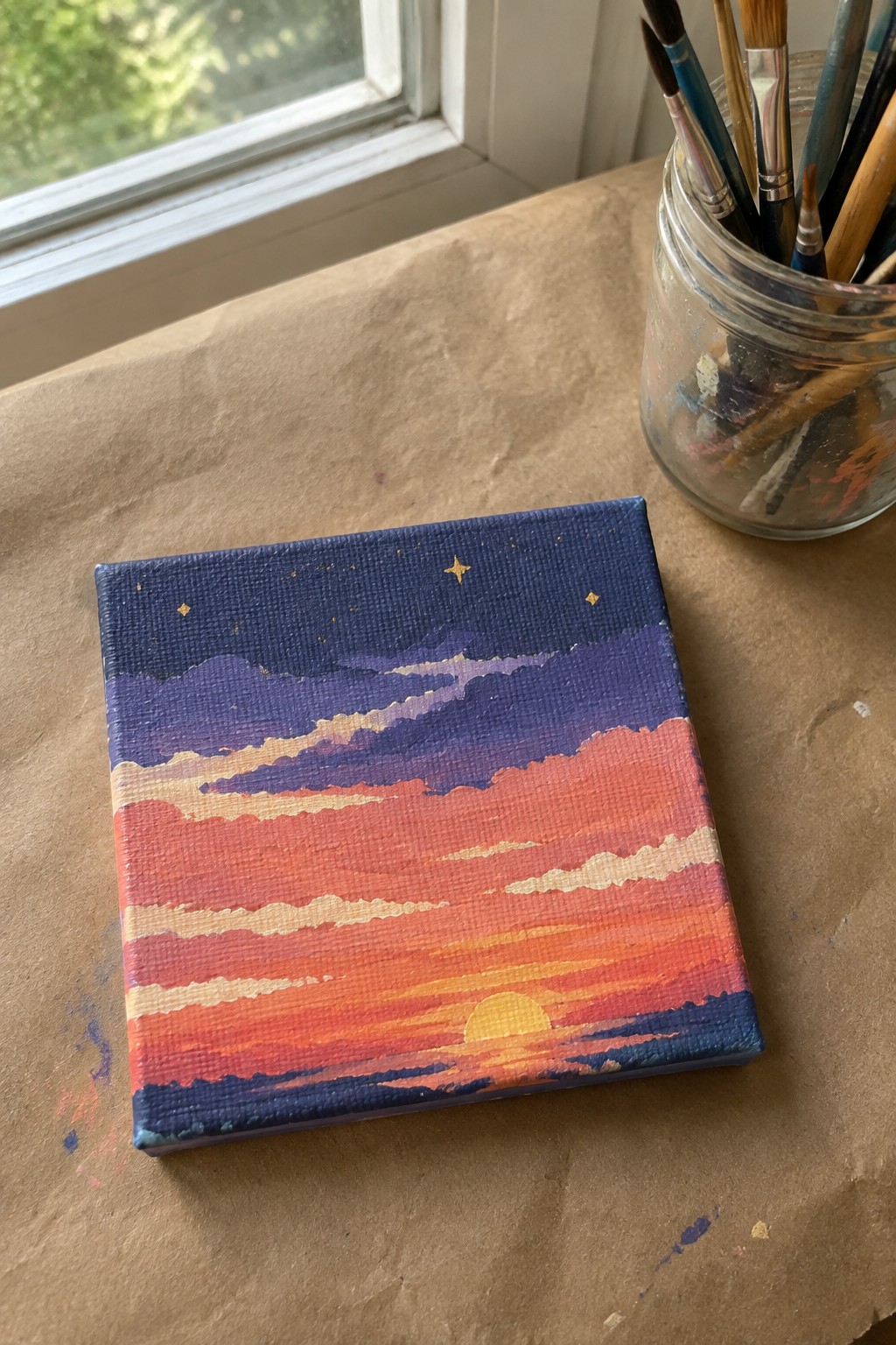 Small canvas sunset painting beside glass jar of paintbrushes on brown paper