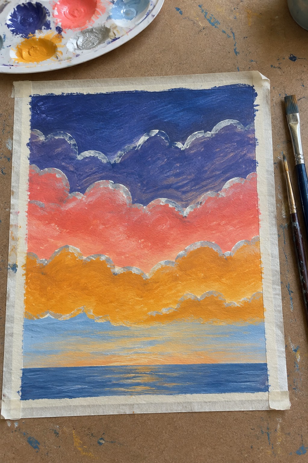 Vibrant sunset painting with layered clouds over water, palette and brushes nearby