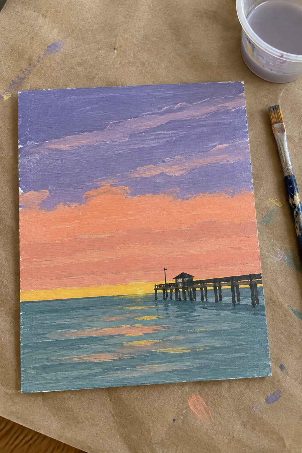 Vibrant sunset painting over ocean with pier, brush and paint cup nearby