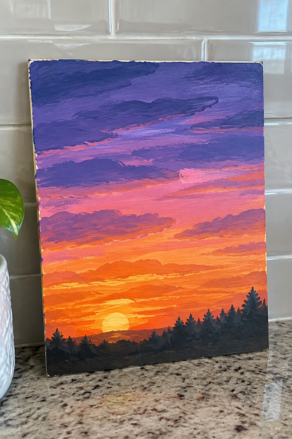 Acrylic painting of a sunset sky with layered purple, pink, and orange clouds above a dark pine tree silhouette.