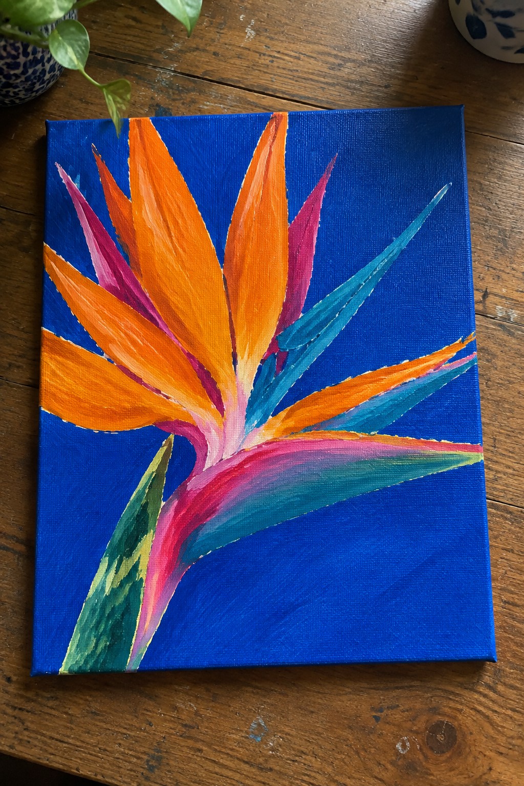 An acrylic painting of a colorful Bird of Paradise flower with orange, pink, and teal petals on a blue canvas.