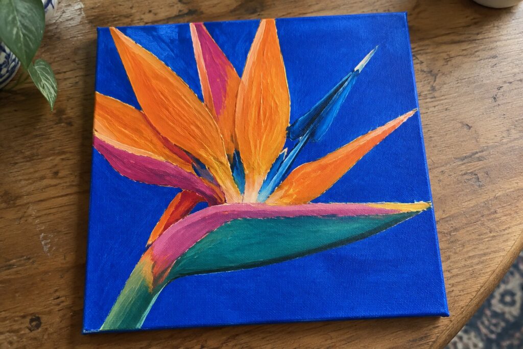 An acrylic painting of a colorful Bird of Paradise flower with orange, pink, and teal petals on a blue canvas.