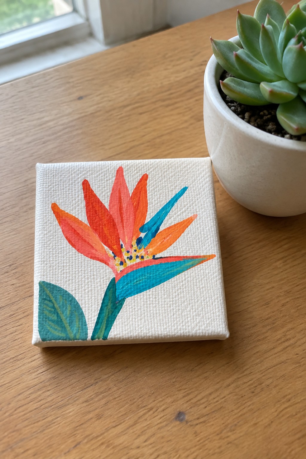 Watercolor bird of paradise painting on small canvas beside potted succulent on wooden table