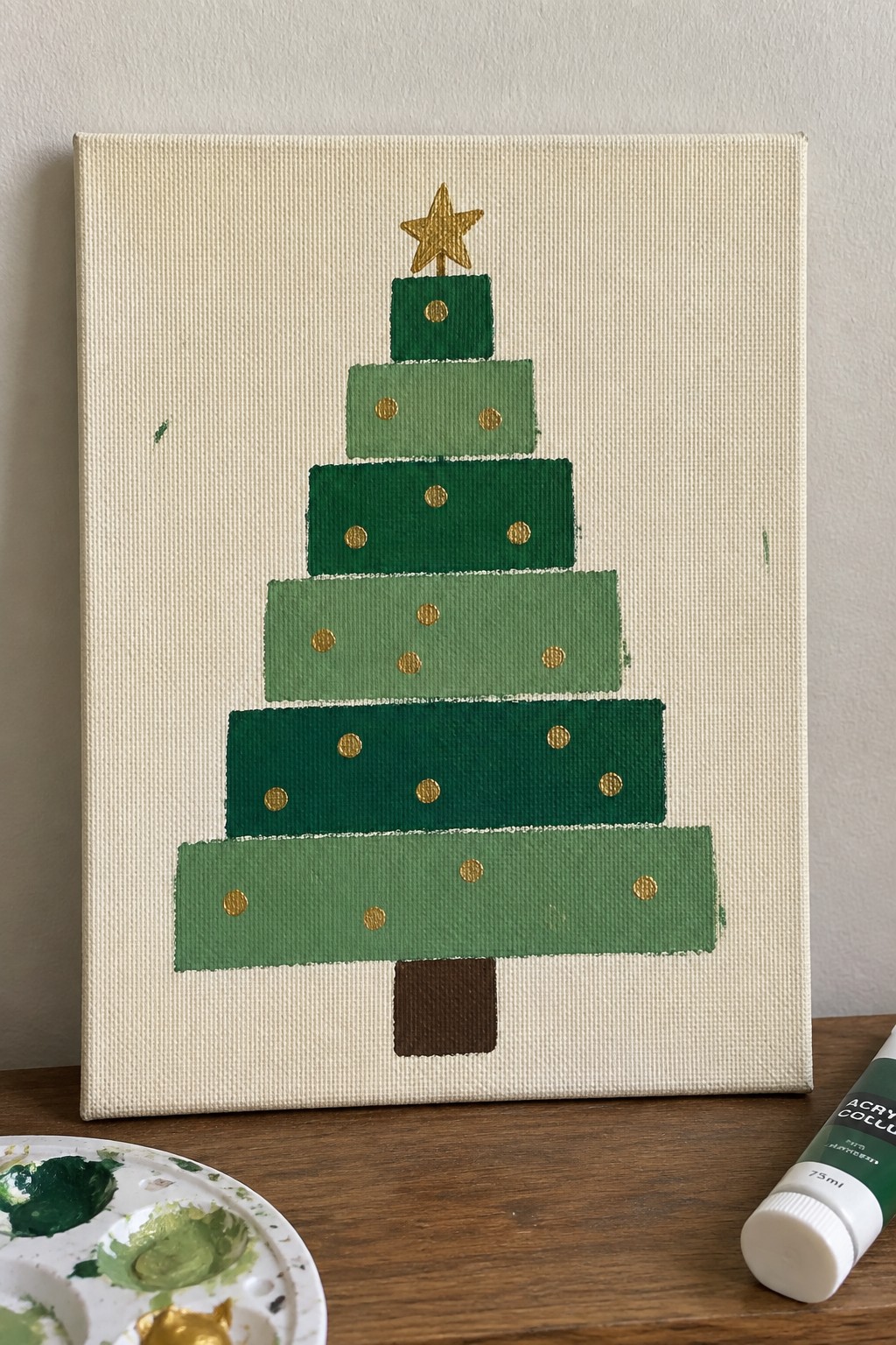 Layered green Christmas tree painting with gold star and dots beside paint palette and tube