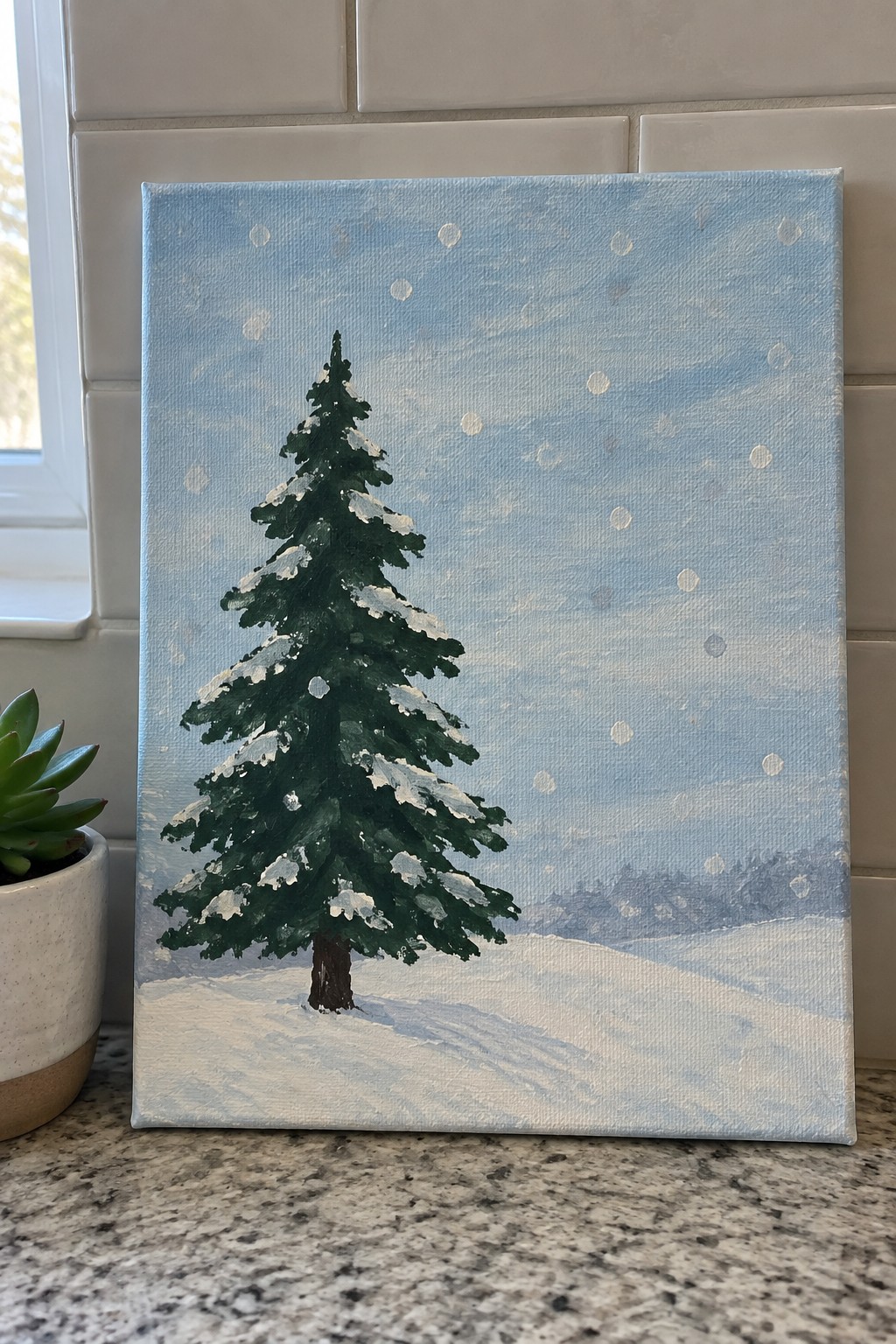Snow-covered pine tree painting on canvas beside potted plant on counter