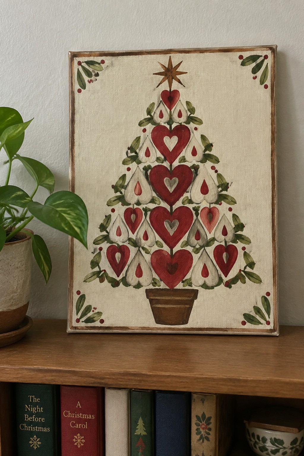 Folk art painting of red heart Christmas tree on wooden shelf with books and plant