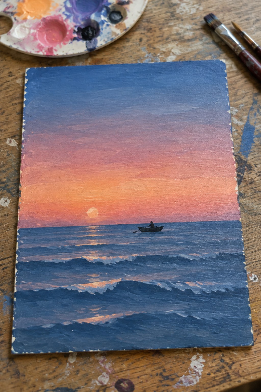 Ocean sunset painting with lone rower in boat, palette and brushes on wood