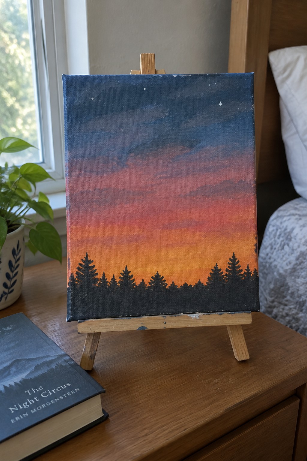 Sunset painting on easel with red-orange sky and pine tree silhouettes beside book and plant.