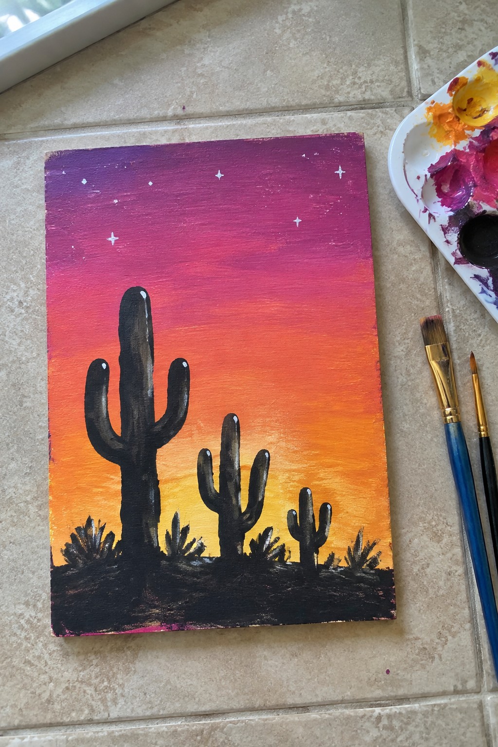 Acrylic painting of silhouetted desert cacti under purple-orange sunset sky with art supplies.
